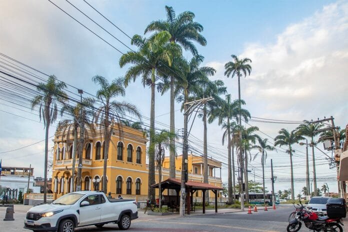 Prédio histórico amarelo do Paço Municipal de Macaé ao lado de fileiras de palmeiras altas, com veículos passando pela rua em primeiro plano. - Fotos : Bruno Moisés