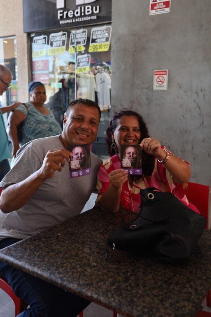 Duas pessoas sentadas em uma mesa sorrindo e segurando panfletos da campanha de combate à violência contra a mulher dentro de uma área comercial.